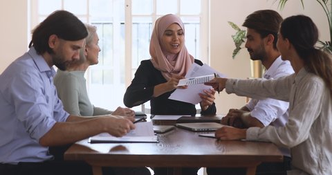 Young female arabian team leader professional in hijab giving out paper marketing research documents to diverse colleagues workers, starting discussing company development strategy in modern office.