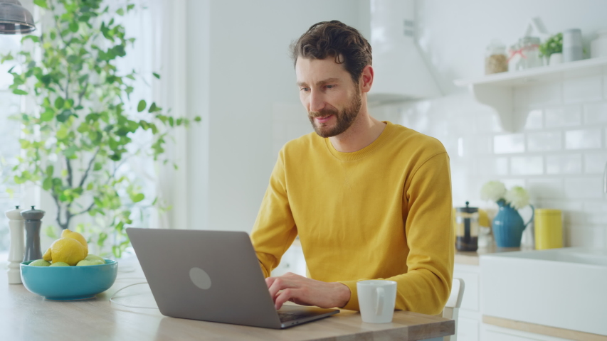 Young Handsome Man Using Laptop Computer and Drinking Morning Cup of Coffee or Tea While Wearing a Yellow Pullover. Curly-hared Male Sitting in a Modern Sunny Kitchen. Freelancer Working from Home. Royalty-Free Stock Footage #1051725616