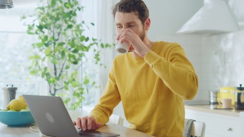 Young Handsome Man Using Laptop Computer and Drinking Morning Cup of Coffee or Tea While Wearing a Yellow Pullover. Curly-hared Male Sitting in a Modern Sunny Kitchen. Freelancer Working from Home.