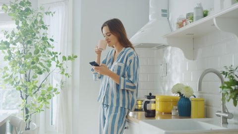 Beautiful Female is Sitting on a Cupboard and Using a Smartphone in a Kitchen While Drinking a Cup of Freshly Brewed Coffee. Girl in Pyjamas with Healthy Lifestyle Relaxes at Home in the Morning.