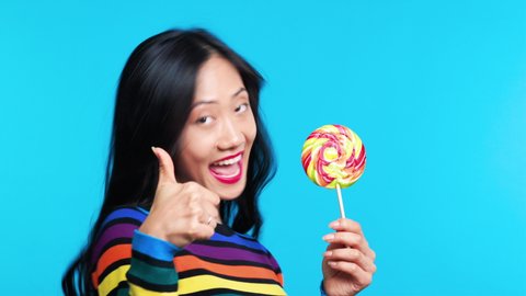 Profile view of young woman licking colorful lollipop on blue background. Tasty food concept