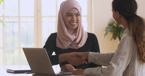Smiling young asian muslim woman in hijab showing presentation on computer to female partner client, shaking hands making agreement, thanking for help, establishing partnership in modern office.