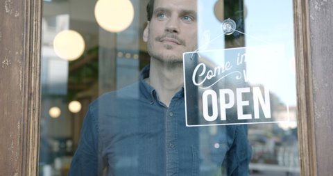 Small Business owner man in apron hanging open sign on door smiling welcoming clients to new cafe restaurant. Entrepreneur Service hospitality.