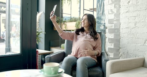 Pregnant woman taking selfie with smart phone in trendy cafe restaurant and sharing with friends pregnancy