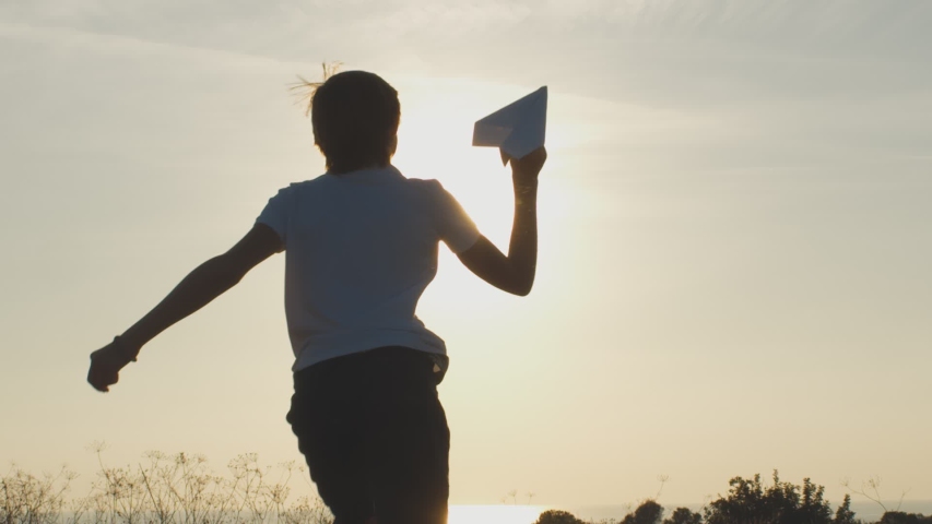 Low angle back view of teen boy running in meadow and launching paper plane against shining sun on sky background. Happy child playing outdoors. Recovery of aviation industry after global crisis