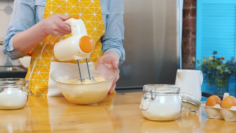 Cropped shot of housewife in yellow apron using mixer machine making dough for bread or cookies. Woman holding glass bowl with dough and electric mixer cooking dessert