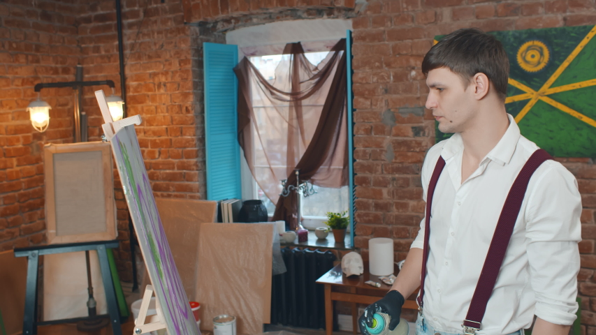 Side view of young male painter in shirt and black gloves using spray paint can working on canvas picture. Street artist painting with spray paint in workshop