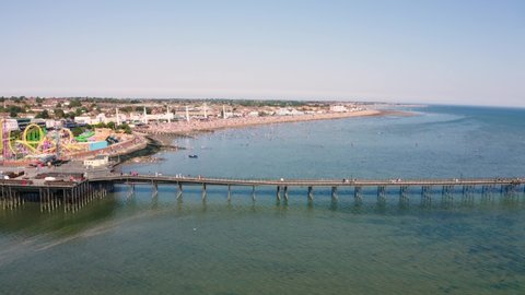 Drone or aerial footage of Southend pier on a busy sunny bank holiday. The beach and sea are full of people and families playing and swimming close to a funfair with a rollercoaster