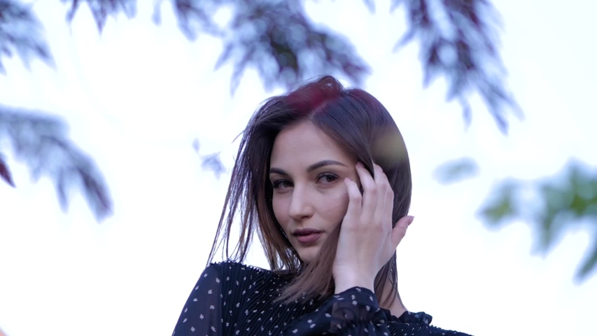 Close-up portrait of cute brunette girl in a black summer dress looking at camera. Slow motion.
