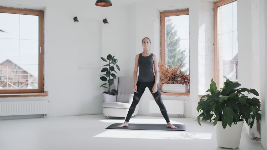 Cameraman shooting fit young brunette girl in sportswear stretching, leaning forward on mat at home. Videographer shoots sporty female doing exercizes, backstage of making video on healthy lifestyle