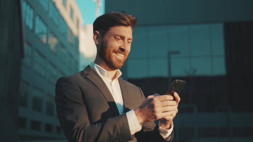 Smiling happy man stand in the city center street uses phone texting scrolling tapping technology communication sunny day success slow motion | Shutterstock HD Video #1051756654