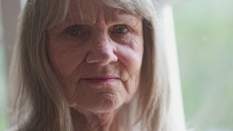 Close up portrait of friendly looking senior Caucasian woman looking at camera. Retired older white woman by a window. Slow motion 4k