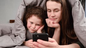 Happy mom with cute kid daughter having fun holding smart phone lying on bed covered with blanket, mother and little girl recording vlog talking to webcam laugh embrace communicate online in internet