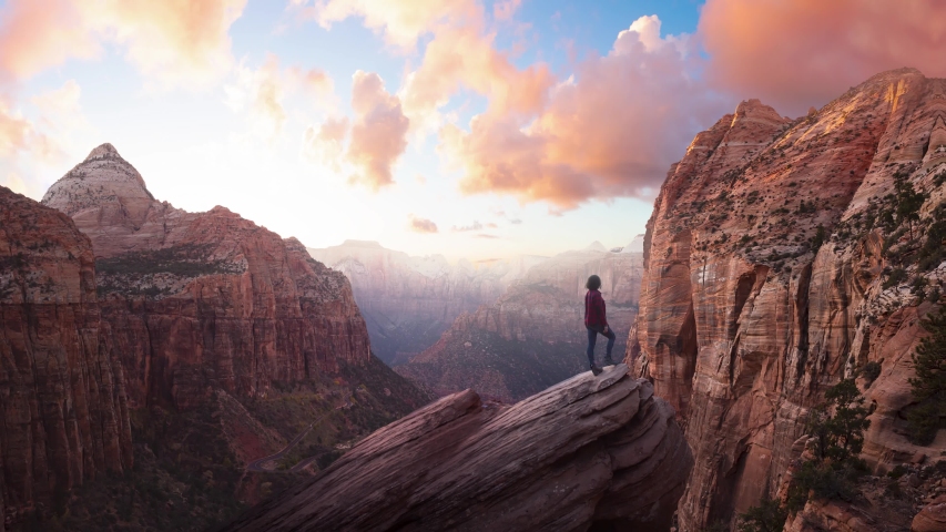 Adventurous Woman at the edge of a cliff is looking at a beautiful landscape view in the Canyon during a vibrant sunset. Taken in Zion National Park, Utah, United States. Parallax Panorama | Shutterstock HD Video #1051773067