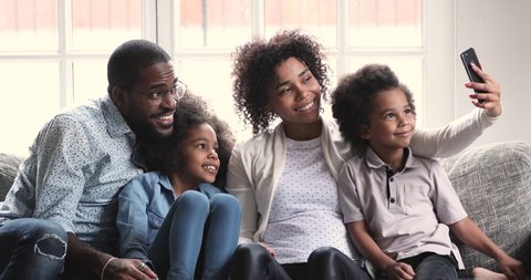 Bonding african american parents sitting on couch with teen daughter and cute preschool son, posing for selfie photo on smartphone. Pretty woman recording mobile video with children and husband.