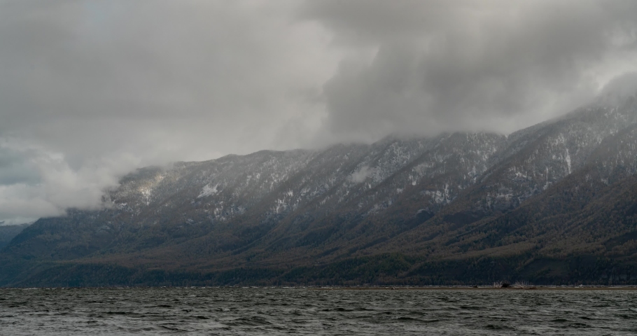 Clouds fly over the snowy mountains and the lake.