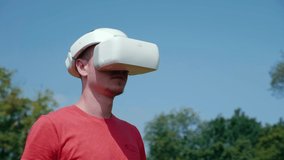 Man in a virtual reality helmet turns his head to the right. The guy in the park in a red T-shirt on a background of blue sky and green trees looks VR glasses. 4K footage