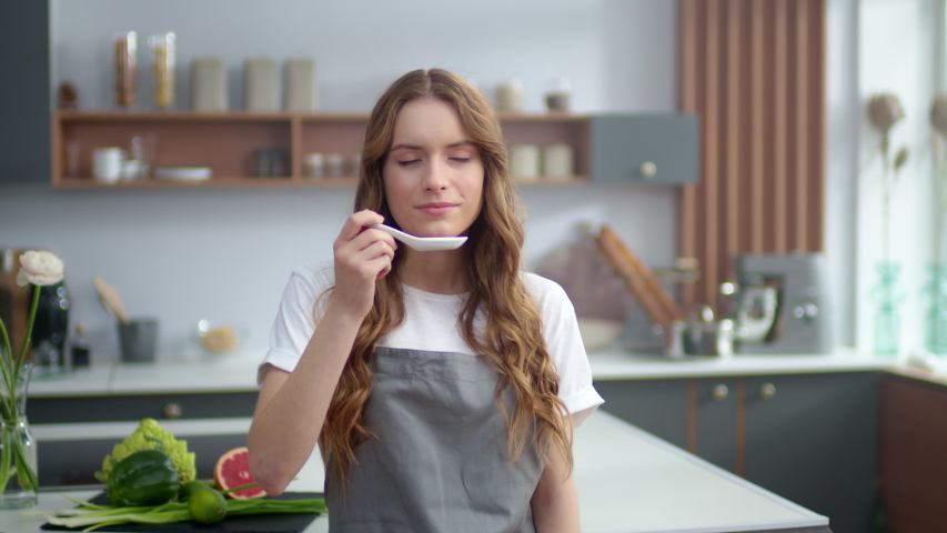 Closeup young woman standing with soup spoon at home kitchen. Cheerful girl smelling soup indoors. Beautiful female chef tasting hot meal in kitchen. Happy woman cooking food indoors. Royalty-Free Stock Footage #1051808563