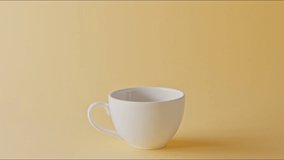 White ceramic cup on a pastel yellow background with fragrant coffee beans pouring in and around on a table. Slow motion, Full HD video, 240fps, 1080p.