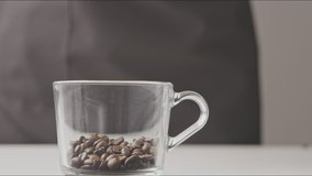 Female's hands fill the glass transparent cup by aromatic roasted coffee beans on a gray background. Slow motion, Full HD video, 240fps, 1080p.
