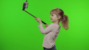 Girl child makes selfie on mobile phone using selfie stick. Kid with pink hair in jeans and pink blouse using smartphone social network app for video call, blogging. Green screen at studio. Chroma key