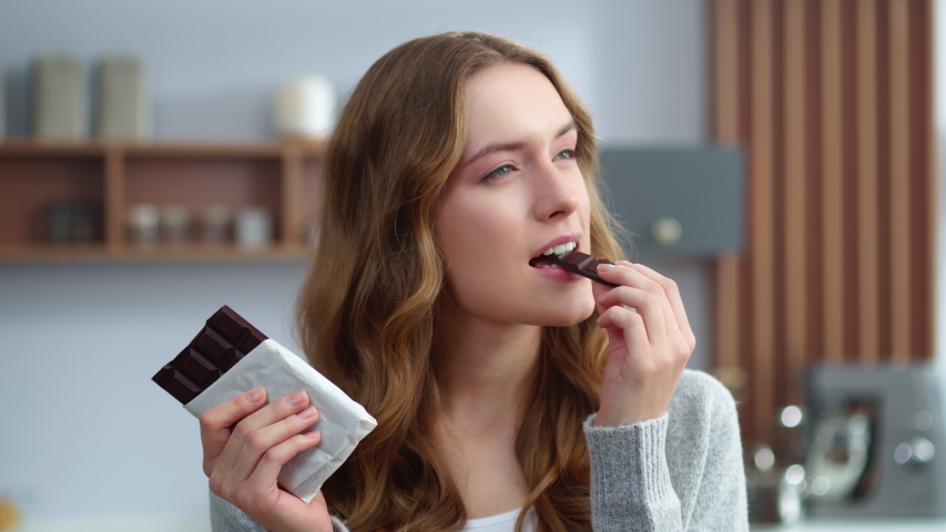 Portrait of young woman eating chocolate at home kitchen. Beautiful female person biting chocolate at modern apartment. Relaxed girl enjoying dark chocolate indoors in slow motion. Royalty-Free Stock Footage #1051823710