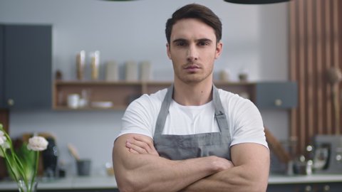 Portrait of serious chef man looking camera at home kitchen. Closeup strict man chef crossing hands indoors. Handsome male professional standing at kitchen background. Young man preparing to cook food