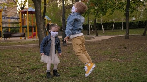 Two cheerful little children in medical masks are playing on the street in the Park, they are jumping on the background of the Playground. Quarantine for the coronavirus. Covid-19 pandemic outbreak.
