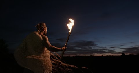 Man in sheepskin with bow and arrows using a flaming branch for light as he walks through the wilderness at night against a sunset sky.