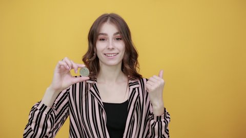Cute Young Woman Light Brown In Striped Pink, Black Shirt On A Yellow Background, Happy Girl Smiling And Holding Coin Bitcoin