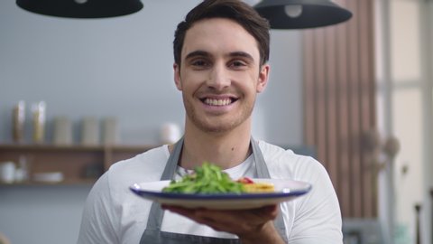 Portrait of smiling chef man holding plate with salad at home kitchen. Cheerful male cook smelling fresh meal at modern apartment. Handsome male professional looking camera at domestic kitchen.
