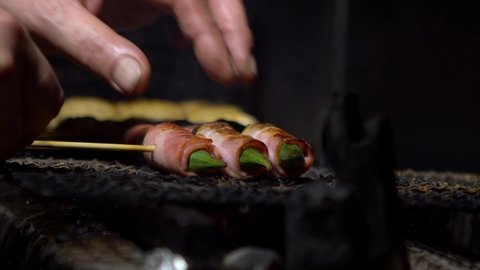 Slow motion of chef cooking pork bacon with okra vegetable inside izakaya Japanese bar in Tokyo. Traditional meat skewers being charcoal grilled in a barbecue. Japan at street food vendor market.-Dan