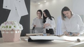 Slow motion video of the three fashion designer works at a table and discuss about something, curves and a pattern, scissors paper, girls arelooking in the smartphone, sew studio, sun light, smiles