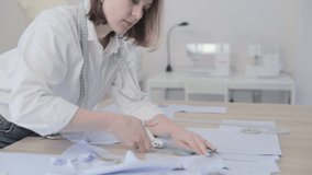 Slow motion video of the fashion designer works at a table with curves and a pattern, she are cutting fabric, around lies scissors, sew machine and dummy on the background, camera is moving back