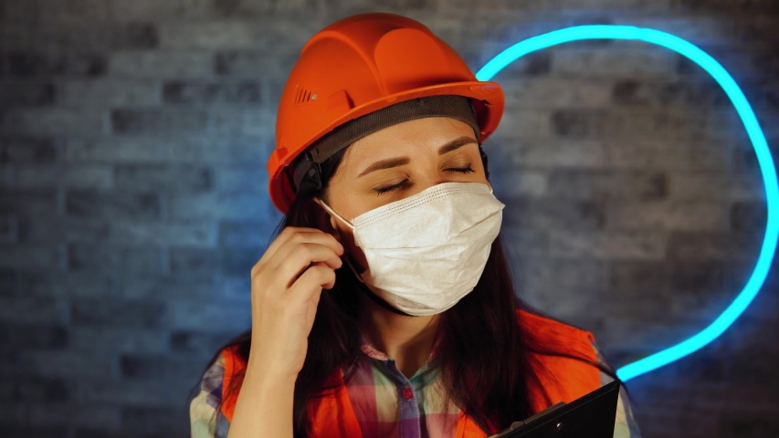 Woman in overalls removing medical mask from face, finishing online work, standing with clipboard against illuminated wall. Concept of threat of coronavirus epidemic infection.