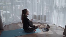 Young calm woman sits in staff easy lotus pose near laptop at home interior, meditating alone on the floor with eyes closed, online yoga training, doing breathing exercise in the morning, no stress