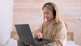 Young girl freelancer works at a laptop at home in her bed. Young woman wears headset conference calling on laptop talks with online teacher studying. Distance learning. Work from home concept.