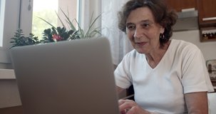 Cheerful senior woman chatting with family online on laptop. Happy elderly female communicating on notebook at home. Hand-held shot, single shot, medium close up, 4K.