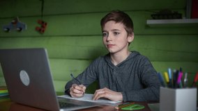 A Schoolboy Studies at Home Remotely Using Video Conferencing. He is Sitting at a Table with a Laptop on a Background of Green Wall. The Boy Greets the Teacher and Writes the Assignment in a Notebook.