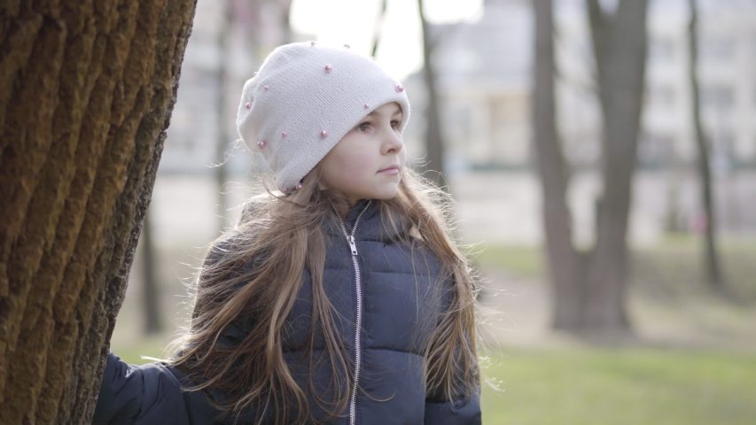 Portrait of surprised Caucasian little girl looking at camera and hiding behind the tree trunk. Joyful kid having fun outdoors on sunny spring day. Nature, lifestyle, relaxation.