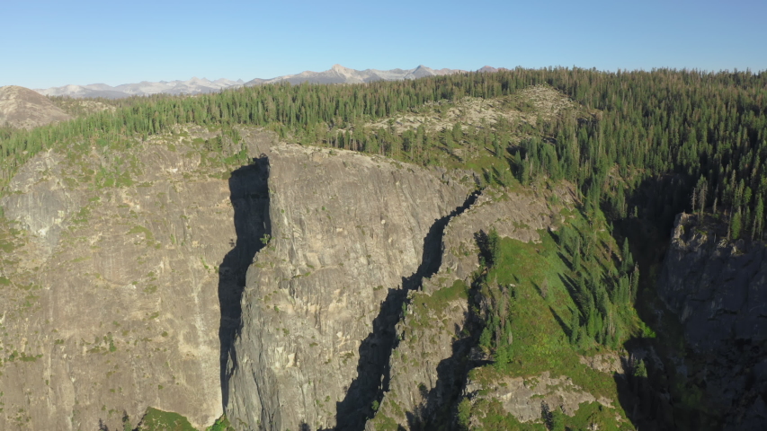 Aerial shot of large granite formations densely covered with conifers under clear blue sky. The top of the mountain is lit with the sun. Yosemite National Park, California, USA. 4K