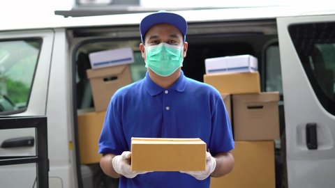 Corona Virus concept, Blue Delivery handsome asian man holding parcel cardboard box with protection mask and medical rubber gloves standing in front of the van. 4k resolution and slow motion shot.