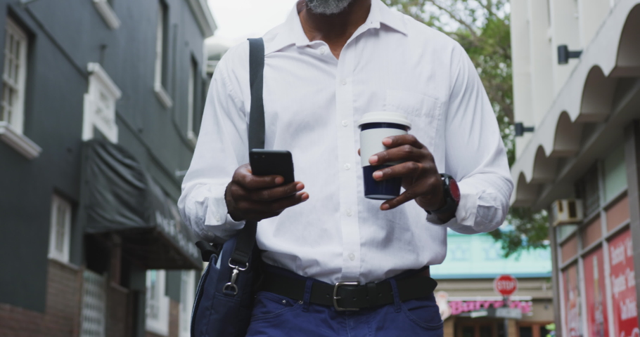 African American man out and about in the city streets during the day, walking down the street, drinking a takeaway coffee and using his smartphone.