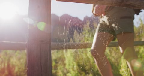 Mid section of Caucasian couple on a trip to the mountains, walking past a fence in a field on a sunny day, a spiderweb in the foreground, in slow motion