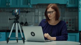 Young 30s female blogger recording video on mobile phone at home on blue kitchen. Work from home woman in glasses talking emotionally to cell camera with laptop in slow motion.