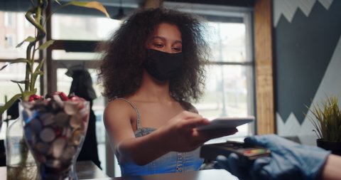 Customer black woman in medical protection mask paying for coffee using NFC technology with phone and credit card, contactless payment with student girl woman after coronavirus quarantine pandemic.