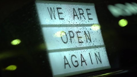 Lightbox with a sign we are open again behind a glass door of the cafe during the rain in the evening. We're open again after quarantine, video of small business owner. Please wear a face mask  