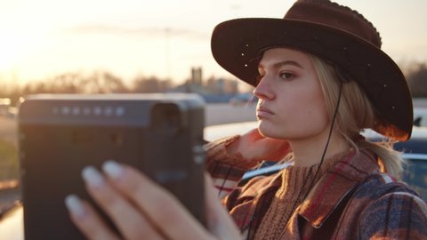 Blonde attractive cowboy girl wearing hat, holding black polaroid camera and preparing for photography. Styled western woman taking pictures outdoors at sunset.