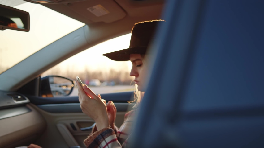 Beautiful western girl wearing cowboy hat reading social media news messaging having rest inside parked car at sunset. Smiling woman. Modern lifestyle.