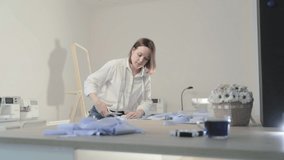 Slow motion video of the fashion designer works at a table with curves and a pattern, she are cutting fabric, around lies scissors, centimeter, sew machine and dummy on the background, Smooth movement